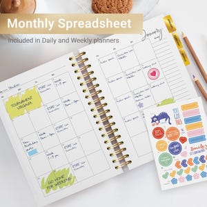 Pu&ograve; includere: Un'agenda mensile con una rilegatura a spirale dorata e una copertina bianca. L'agenda ha una griglia di calendario con note scritte a mano e note adesive colorate. Le note includono "Tournament Virginia", "Work 9am-3pm", "ESRE Lab Chemistry", "Den Birthday Party", "Work 1-9pm", "Go Home for Weekend", "Public Speech", "Chemistry Lab Math", "Cooking Club", "Return Books" e "January". L'agenda include anche un foglio di adesivi colorati con vari disegni, tra cui un gatto, un cuore e frecce.