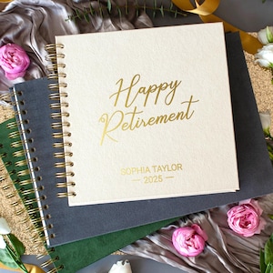 May include: A cream-colored spiral-bound notebook with the words "Happy Retirement" in gold script. The name "Sophia Taylor" and the year "2025" are printed below. The notebook rests on top of a dark gray and green notebook, with pink roses and gold ribbon accents.