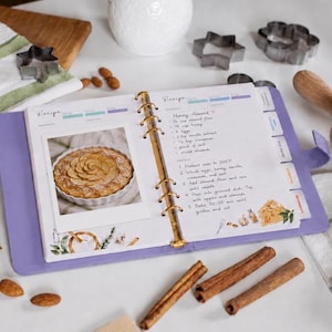 May include: A lavender-colored ring-bound recipe book open to a page with a handwritten Honey Almond recipe and a photo of a tart. The page features watercolor illustrations of kitchen tools and ingredients, with cinnamon sticks and almonds nearby.