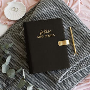 May include: A black notebook with the gold text future MRS. JONES, featuring a gold metal clasp closure, accompanied by a slim gold pen, resting on a grey textured knit fabric with a white shell-shaped dish containing two silver rings.