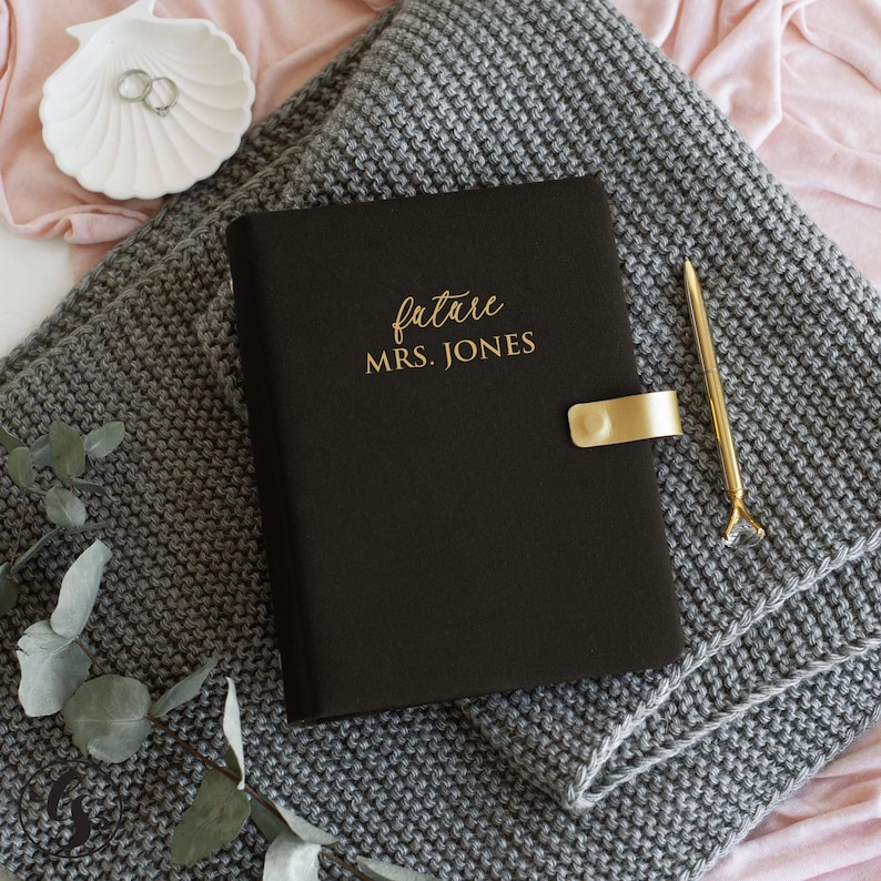 May include: A black notebook with the gold text future MRS. JONES, featuring a gold metal clasp closure, accompanied by a slim gold pen, resting on a grey textured knit fabric with a white shell-shaped dish containing two silver rings.