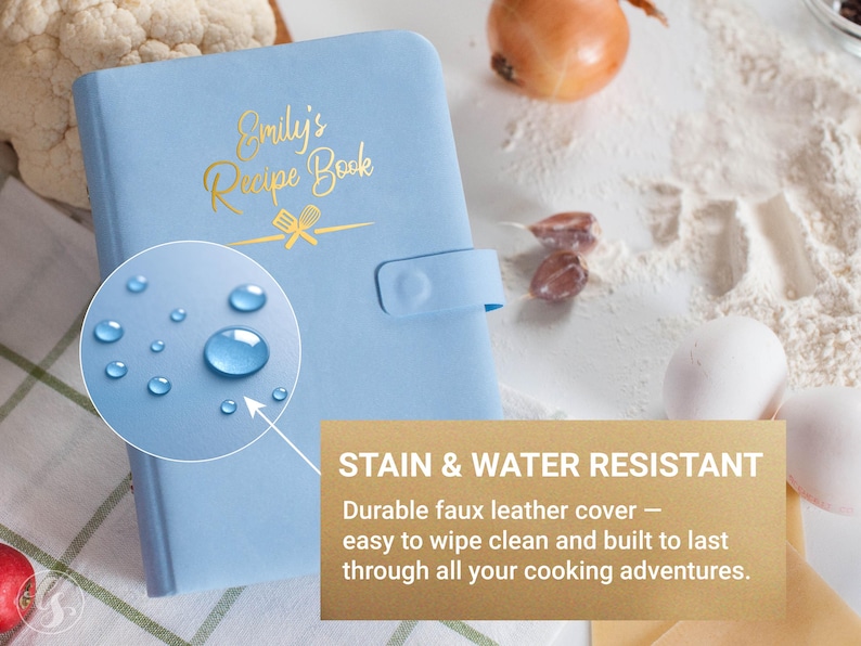 May include: A light blue recipe book with the text "Emily's Recipe Book" in gold lettering. The book has a snap closure and is stain and water resistant. The image also shows water droplets.