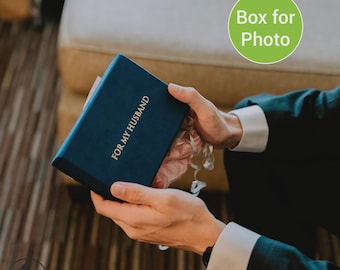 May include: A navy blue box with the text "FOR MY HUSBAND" in gold lettering. The box is being opened, revealing a pink interior and silver ribbon. The box is held by a person wearing a dark suit with white cuff links.