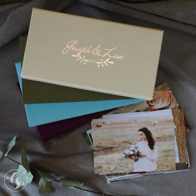 May include: A stack of four photo albums with a beige top album that has the text "Joseph & Lisa" in gold lettering. The top album is closed and the other three albums are open.