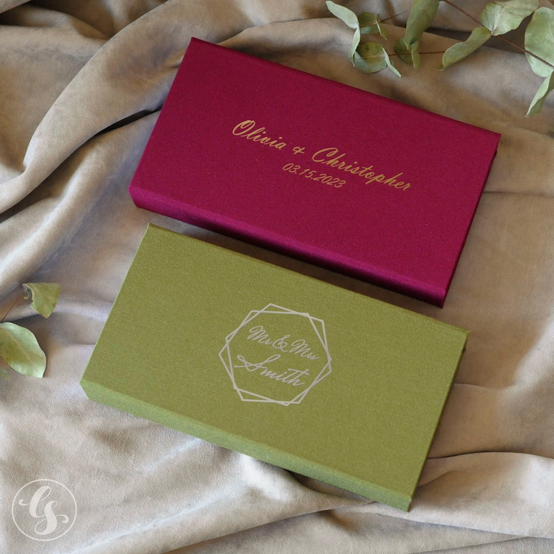 May include: Two rectangular boxes with personalized engravings. The top box is burgundy with gold lettering that reads "Olivia & Christopher 03.15.2023". The bottom box is green with a silver geometric design and the text "Mr. & Mrs. Smith".