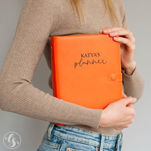 Puede incluir: Agenda de color naranja brillante con el texto "KATYA'S planner" en escritura negra. El planificador tiene un cierre de broche. Una persona lleva un suéter color beige y vaqueros azules.