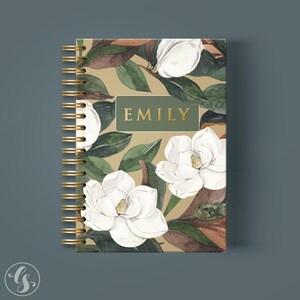May include: A spiral-bound notebook featuring a tan background with a watercolor pattern of white magnolia flowers and dark green leaves. A central rectangular green label displays the name EMILY in gold capital letters on the front cover.
