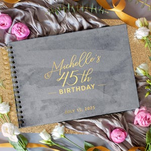 May include: A gray spiral-bound notebook with gold lettering that reads "Michelle's 45th Birthday" and the date "July 15, 2025". The notebook is surrounded by flowers, ribbons, and a gold glitter surface.