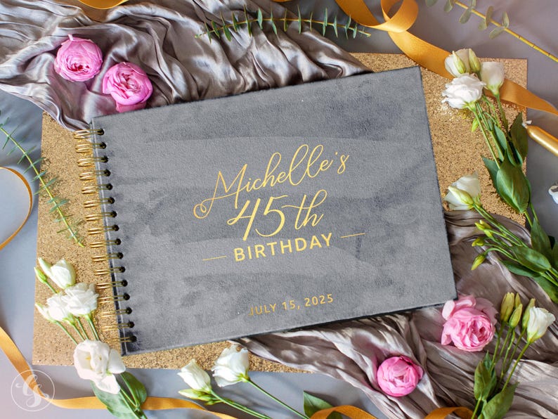May include: A gray spiral-bound notebook with gold lettering that reads "Michelle's 45th Birthday" and the date "July 15, 2025". The notebook is surrounded by flowers, ribbons, and a gold glitter surface.