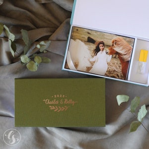 May include: A green box with a gold inscription that reads "2023. Charles & Betty" with a gold leaf design. The box is open and contains a photo album with a photo of a woman in a white dress with a horse.
