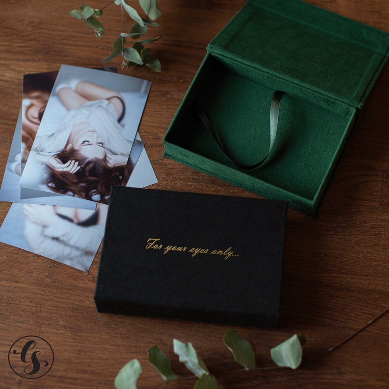 K&ouml;nnte beinhalten: Eine schwarze Schachtel mit goldenem Text "For your eyes only..." liegt auf einer Holzoberfl&auml;che neben gedruckten Fotos und einer offenen gr&uuml;nen Samtbox. Die gr&uuml;ne Box hat ein Band im Inneren. Eukalyptusbl&auml;tter sind um die Gegenst&auml;nde verstreut.