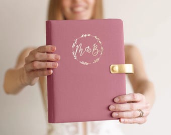 May include: A dusty rose fabric-covered journal featuring a gold foil wreath design on the front cover with the letters M and B centered inside. The journal includes a gold metal snap closure on the right side to secure the cover shut.