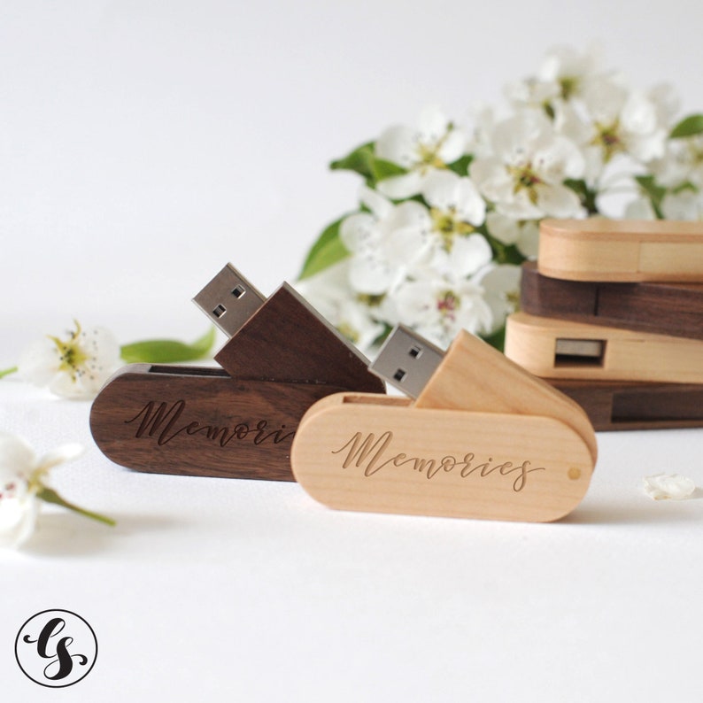 Puede incluir: Dos memorias USB de madera con la palabra "Memories" grabada en ellas. Las memorias est&aacute;n abiertas y los conectores USB son visibles. Las memorias est&aacute;n sobre una superficie blanca con flores blancas en el fondo.