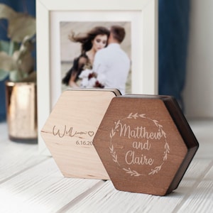May include: Two hexagonal wooden ring boxes with engraved text. The box on the left is light wood with the text "William 6.16.20". The box on the right is dark wood with the text "Matthew and Claire" in a wreath.