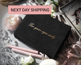 May include: A black velvet box with gold lettering that reads "For your eyes only". The box is sitting on a table with a white flower and a piece of lace.