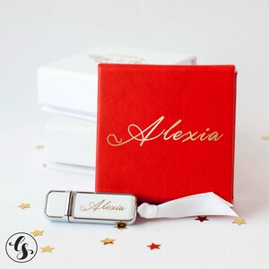 K&ouml;nnte beinhalten: Ein wei&szlig;er USB-Stick mit dem Namen "Alexia" in goldenen Lettern graviert. Der Stick befindet sich in einer roten Geschenkbox mit dem gleichen Namen in goldenen Lettern graviert.