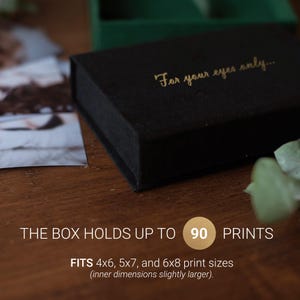 K&ouml;nnte beinhalten: Eine schwarze Box mit dem goldenen Text "For your eyes only...". Die Box fasst bis zu 90 Abz&uuml;ge und passt f&uuml;r Druckgr&ouml;&szlig;en von 10x15 cm, 13x18 cm und 15x20 cm. Die Box steht auf einer Holzoberfl&auml;che.