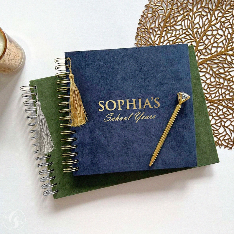 May include: Two spiral-bound journals, one green and one navy blue, with gold lettering that reads "SOPHIA'S School Years." A gold pen with a diamond-shaped top rests on the navy journal. A gold tassel hangs from the navy journal, and a silver tassel hangs from the green journal.