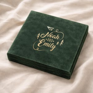 May include: A square, forest green velvet box with the names "Noah and Emily" in gold script. A paper airplane and two small trees are also in gold. The box is on a cream-colored fabric surface.