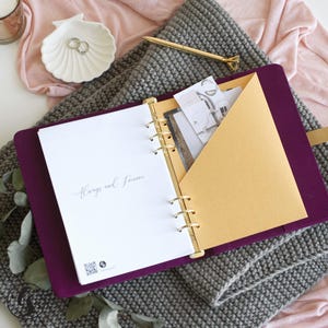 May include: A deep purple ring-bound planner with gold hardware, featuring a white page with the text Always and Forever, a gold interior pocket containing paper inserts, a gold pen, a white shell-shaped dish, and a gray textured fabric.