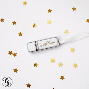 K&ouml;nnte beinhalten: Ein silberner und wei&szlig;er USB-Stick mit dem Namen "Alexia" in goldenen Lettern graviert. Der Stick ist von goldenen Sternen auf wei&szlig;em Hintergrund umgeben.