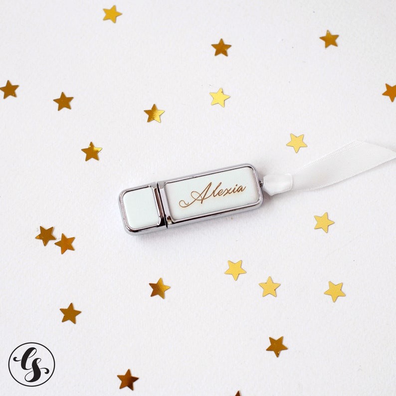 K&ouml;nnte beinhalten: Ein silberner und wei&szlig;er USB-Stick mit dem Namen "Alexia" in goldenen Lettern graviert. Der Stick ist von goldenen Sternen auf wei&szlig;em Hintergrund umgeben.