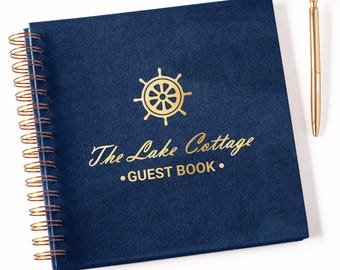 May include: A square navy blue guest book with gold spiral binding. The cover features a gold ship wheel icon and the text "The Lake Cottage GUEST BOOK" in gold lettering. A gold pen with a clear diamond-shaped topper is positioned to the right.