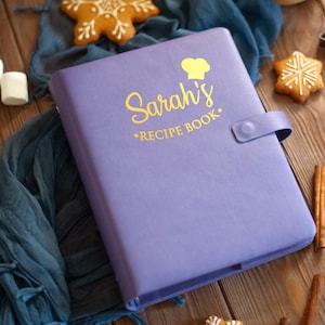 May include: A purple recipe book with a snap closure featuring a yellow chef hat icon and the text Sarah's RECIPE BOOK printed on the front cover. The book has a smooth, matte finish and is positioned at an angle with a visible spine and strap.