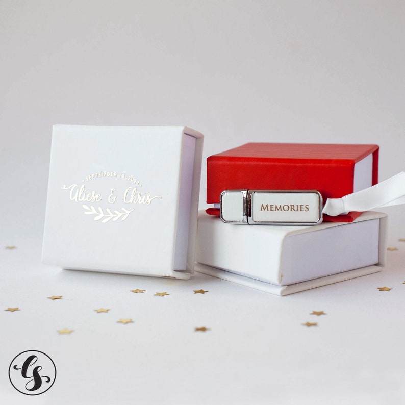 K&ouml;nnte beinhalten: Zwei wei&szlig;e Geschenkboxen mit einem silbernen USB-Stick in der Mitte. Der USB-Stick tr&auml;gt die Aufschrift "Memories". Die obere Box ist rot. Die untere Box tr&auml;gt die Aufschrift "September 14, 2021, Alise & Chris".