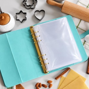 May include: A light blue A5 binder with a gold-colored six-ring metal mechanism and a clear plastic pocket insert. The binder is open, revealing a smooth interior surface, and is surrounded by various kitchen items including cookie cutters.