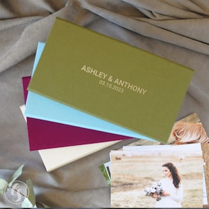 May include: A stack of four photo albums in different colors, including green, blue, burgundy, and cream. The top album has the text "ASHLEY & ANTHONY 03.15.2023" printed on the front.