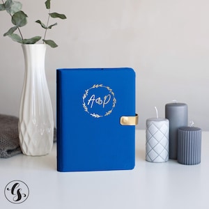 May include: A vibrant royal blue notebook featuring a gold foil wreath design on the cover with the letters A and P and a ring icon in the center. The notebook has a gold metal clasp closure and is positioned upright on a white surface.