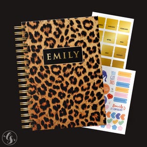 May include: A leopard print spiral bound notebook with the name "EMILY" printed in gold on the front cover. The notebook comes with a set of gold stickers with months of the year and a set of colourful stickers with motivational phrases and designs.