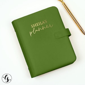 May include: A sage green planner with a gold-toned closure strap and the text SHEILA'S planner printed in gold script on the front cover, accompanied by a slim gold-toned metal pen positioned diagonally in the upper right corner.