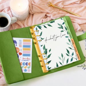 May include: A green ring-bound planner with gold hardware, featuring a white page with green leaf illustrations and the text "do what you love". A sticker sheet with various colorful icons and the text "YOU GOT THIS" and "Smile, IT'S CONTAGIOUS" is tucked into the inside cover.