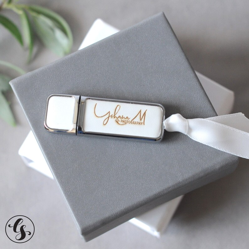 K&ouml;nnte beinhalten: Ein silberner USB-Stick mit einem wei&szlig;en Etikett, auf dem "Johanna M Photography" in goldenen Buchstaben steht. Der Stick liegt auf einer grauen Box.