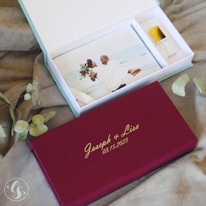 May include: A burgundy velvet box with gold lettering that reads "Joseph & Lisa 03.15.2023". The box is open and contains a white box with a photo of a couple and a small bottle.