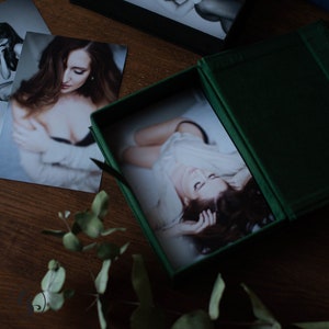 K&ouml;nnte beinhalten: Eine dunkelgr&uuml;ne Samtfotobox mit einem Foto darin. Mehrere Fotos sind auf einer Holzoberfl&auml;che um die Box herum verstreut. Die Fotos zeigen eine Frau in verschiedenen Posen. Eukalyptusbl&auml;tter befinden sich im Vordergrund.