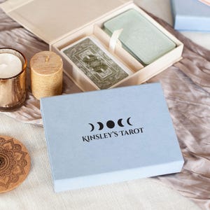 Könnte beinhalten: Eine hellblaue rechteckige Schachtel mit der Aufschrift KINSLEY'S TAROT und einem Mondphasen-Symbol, neben einer offenen Schachtel mit zwei Kartendecks, einem metallischen Kerzenhalter mit weißer Kerze, einer strukturierten goldenen Stumpenkerze und einem runden Holzuntersetzer.