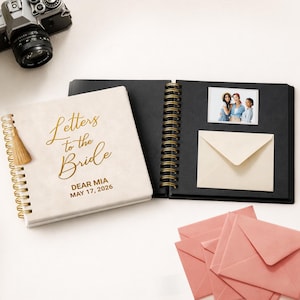 May include: A square spiral-bound scrapbook with a cream cover featuring gold text that reads Letters to the Bride, DEAR MIA, MAY 17, 2026. The open book displays black pages with a photo of three people and a cream envelope, plus pink envelopes.
