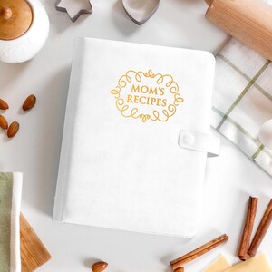 May include: A white recipe binder featuring a gold-colored decorative frame and the text "MOM'S RECIPES" printed on the front cover. The binder includes a side tab with a snap closure and has a smooth, solid white finish on the exterior cover.