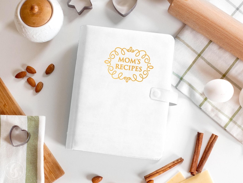 May include: A white recipe binder featuring a gold-colored decorative frame and the text "MOM'S RECIPES" printed on the front cover. The binder includes a side tab with a snap closure and has a smooth, solid white finish on the exterior cover.
