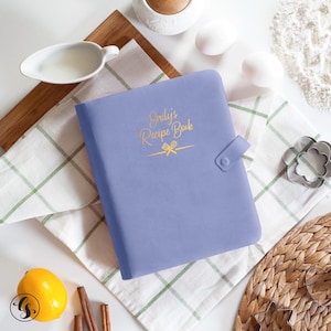 May include: A periwinkle-colored recipe book with the text "Emily's Recipe Book" in gold script. The book has a snap closure and is surrounded by baking ingredients, including eggs, a lemon, and cinnamon sticks.