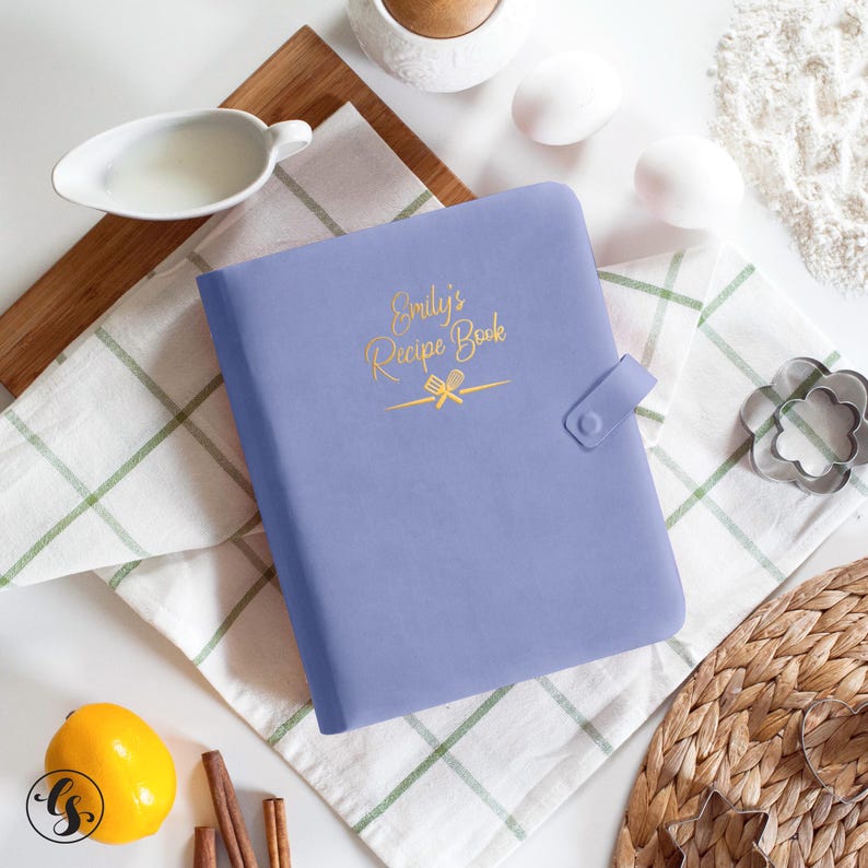 May include: A periwinkle-colored recipe book with the text "Emily's Recipe Book" in gold script. The book has a snap closure and is surrounded by baking ingredients, including eggs, a lemon, and cinnamon sticks.