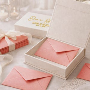 May include: A cream-coloured hinged keepsake box containing a coral-pink envelope, with a matching box lid nearby featuring gold text that reads Dear One LETTERS TO THE BRIDE MAY 15, 2026, alongside a stack of coral-pink envelopes tied with a cream ribbon.
