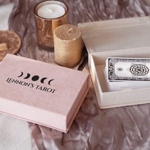 May include: A pink box with the text "Lennon's Tarot" and a moon symbol. The box is open and shows a deck of tarot cards in a white box.