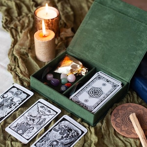 Könnte beinhalten: Eine grüne Samtbox mit einem Scharnierdeckel, die ein Tarotkartenset, eine kleine Kristalltasche und eine kleine Kräuterbeutel enthält. Die Box steht auf einer grünen Stoffoberfläche mit zwei Kerzen.