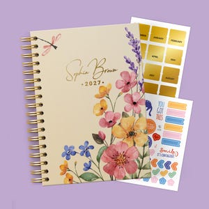 May include: A spiral-bound planner with a cream cover featuring watercolour flowers, a dragonfly, and the text Sophia Brown 2027. Included are two sticker sheets with gold month labels and various colourful icons, shapes, and motivational phrases.