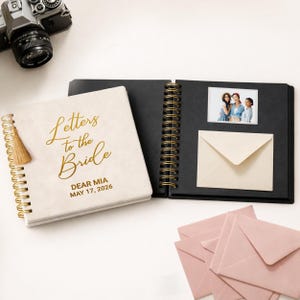 May include: A cream-colored spiral-bound notebook with gold lettering that reads "Letters to the Bride" and "Dear Mia May 17, 2026." The notebook is next to an open black photo album with a photo and an envelope. Pink envelopes are also visible.