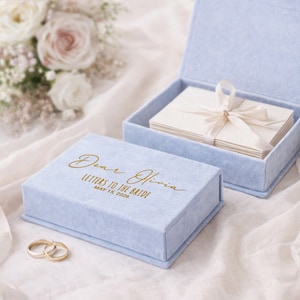 May include: A light blue velvet box with gold lettering that reads "Dear Olivia, Letters to the Bride, May 15, 2026." Inside the open box are white cards tied with a cream ribbon. Two gold wedding rings are in the foreground.